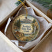 A decorative ornament in a box, featuring a scenic landscape with mountains, pine trees, and a waterfall.