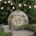 A round ornament with a lighthouse and beach scene, featuring a bird, turtle, and palm tree, is displayed on a table with a blurred background of greenery and lights.