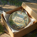 A decorative ornament in a wooden box, featuring a scenic landscape with mountains, trees, and wildlife.