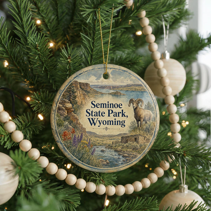 A round ornament hanging on a Christmas tree, featuring a scenic landscape with a river and mountains, and a goat in the foreground.