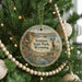 A round ornament hanging on a Christmas tree, featuring a scenic landscape with a river and mountains, and a goat in the foreground.