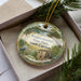 A decorative ornament in a box, featuring a scenic landscape with a house, trees, and a bird.
