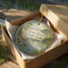 A circular ornament with a scenic landscape of a river and trees, placed in a wooden box with a gold string.