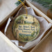 A round ornament with a picture of a man and text, placed in a box with a white tissue paper lining.