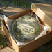 A decorative ornament in a wooden box, featuring a scenic landscape with a river, mountains, and wildlife.