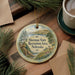 A round ornament with a scenic landscape of Sherman State Recreation Area, Nebraska, featuring a deer and pine trees, is displayed on a wooden surface alongside a cup of coffee and some papers.