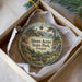 A round ornament with a scenic landscape of a coastal area, featuring a lighthouse, trees, and water, is displayed in a box.