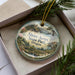A decorative ornament in a box, featuring a scenic landscape with mountains, trees, and water.