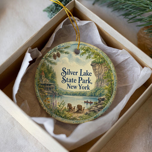 A round ornament in a box, featuring a scenic landscape with a lake, mountains, and a cabin.