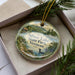 A decorative ornament in a box, featuring a scenic landscape with a lake, mountains, and pine trees.