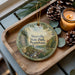 A decorative ornament with a scenic landscape of Sizersville State Park, Pennsylvania, featuring a river, forest, and mountains. The ornament is placed on a wooden tray, with pine cones and a lit candle nearby.