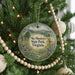 A round ornament hanging on a Christmas tree, featuring a scenic landscape with mountains, trees, and wildlife.