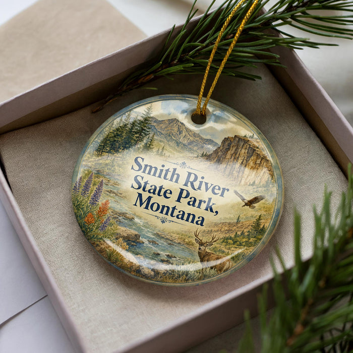 A decorative ornament in a box, featuring a scenic landscape with mountains, pine trees, and wildlife.