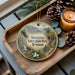 A wooden tray with a decorative ornament featuring a scenic mountain landscape and pine cones, along with a lit candle and pine cones.