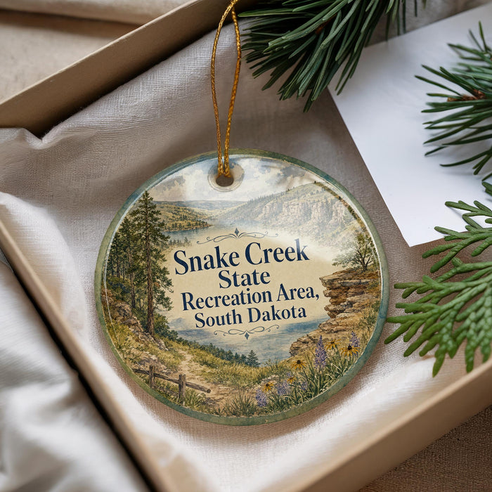 A decorative ornament with a scenic landscape of Snake Creek State Recreation Area in South Dakota, featuring a river, mountains, and pine trees.