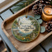 A decorative ornament featuring a lighthouse and a scenic landscape, placed on a wooden tray alongside pine cones and a lit candle.