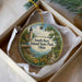 A round ornament with a lighthouse and a scenic landscape, placed in a box with a white tissue paper lining.