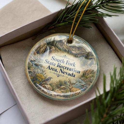 A decorative ornament with a landscape scene featuring mountains, pine trees, and wildlife, including a bird and a deer, is displayed in a box.