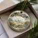 A decorative ornament with a landscape scene featuring mountains, pine trees, and wildlife, including a bird and a deer, is displayed in a box.