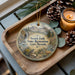 A decorative ornament with a scenic landscape featuring a waterfall, pine trees, and a deer, placed on a wooden tray alongside pine cones and a lit candle.