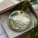 A decorative ornament in a box, featuring a scenic landscape with a beach, pine trees, and a body of water.