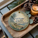A decorative ornament with a scenic image of South Whidbey State Park, Washington, sits on a wooden tray alongside pine cones and a lit candle.