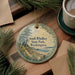 A round ornament with a scenic image of South Whidbey State Park, Washington, hanging on a string. The ornament is placed on a wooden surface, surrounded by pine branches and a cup of coffee.