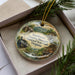 A decorative ornament in a box, featuring a scenic landscape with a train, buildings, and greenery.