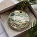 A decorative ornament in a box, featuring a scenic landscape with a lighthouse and pine trees.