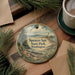 A decorative ornament with a lighthouse and a scenic landscape, featuring the text "Spenser Spit State Park, Washington" on it, is displayed on a wooden surface alongside a cup of coffee and some pine branches.