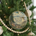 A round ornament hanging on a Christmas tree, featuring a scenic landscape with a lighthouse and a beach.
