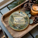 A decorative ornament with a scenic landscape of Spring Creek State Recreation Area, South Dakota, featuring pine cones, a candle, and greenery.