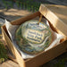 A decorative ornament in a wooden box, featuring a scenic landscape with mountains, waterfalls, and pine trees.