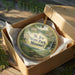 A circular ornament in a box, featuring a scenic landscape with a waterfall and a house, with the text "St. Joe State Park, Missouri" printed on it.