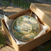 A round ornament in a box, featuring a scenic landscape with mountains, trees, and a lake, with the text "Steinaker State Park, Utah" printed on it.