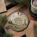 A round ornament with a scenic landscape of Steptoe Battlefield State Park, Washington, featuring pine trees and a monument, is displayed on a wooden surface alongside a cup of coffee and some papers.