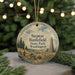 A round ornament with a scenic landscape featuring a tall monument, trees, and a fence, hanging in front of a Christmas tree with lights.
