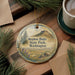 A decorative ornament featuring a scenic landscape with mountains, pine trees, and a body of water, hanging on a wooden surface.