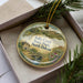 A decorative ornament in a box, featuring a scenic landscape with a house, bridge, and flowers.