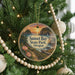 A round ornament hanging on a Christmas tree, featuring a scenic landscape with a sunset over a body of water and a forested area.