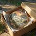 A decorative ornament in a box, featuring a scenic landscape with a sunset over a body of water and mountains in the background.