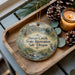 A wooden tray with a decorative ornament featuring a scenic landscape and a bird, along with pine cones and a lit candle, creating a cozy and rustic atmosphere.