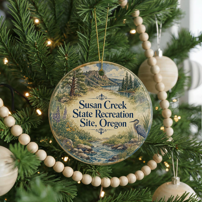 A round ornament hanging on a Christmas tree, featuring a scenic landscape with a river, trees, and a bird.