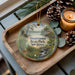 A decorative ornament featuring a scenic landscape of Swartswood State Park, New Jersey, with pine cones and a lit candle on a wooden tray.