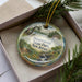 A decorative ornament in a box, featuring a scenic landscape with a river, trees, and a house, with the text "Swartswood State Park, New Jersey" printed on it.