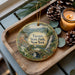 A decorative ornament featuring a scenic landscape of Taconic State Park, New York, with pine cones, a candle, and greenery surrounding it.