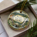 A decorative ornament in a box, featuring a scenic landscape with a waterfall and pine trees.