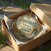 A decorative ornament in a box, featuring a scenic landscape with mountains, trees, and a waterfall.