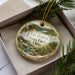 A decorative ornament with a scenic landscape of a waterfall and forest, placed in a box with pine branches.