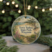 A round ornament with a painting of a park scene, featuring a bird, trees, and a body of water, hanging in front of a Christmas tree with lights.